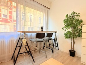 Une pièce avec une table en bois et deux chaises, une plante en pot, et une vue sur des immeubles à travers une fenêtre ornée de rideaux légers.