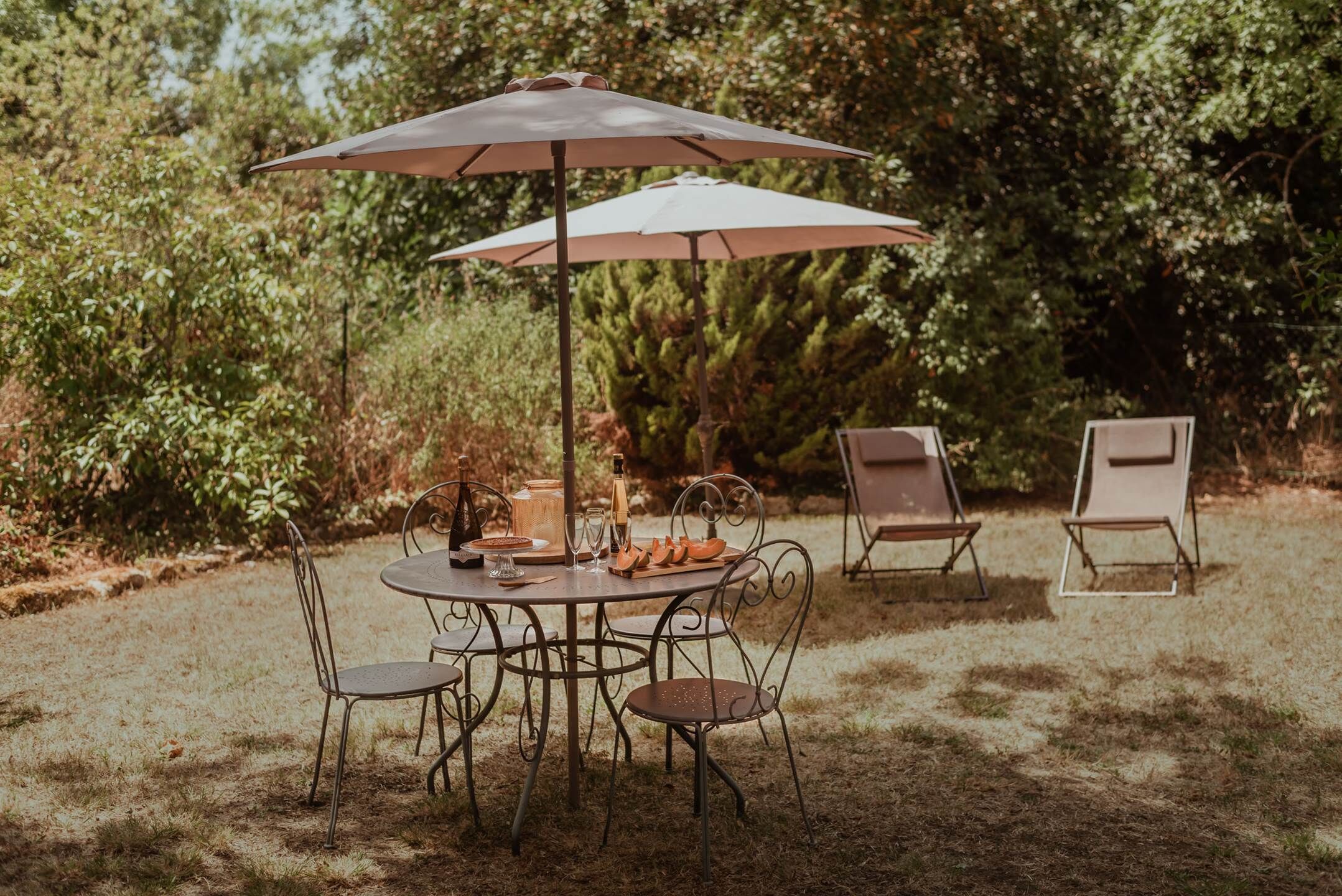 Un área de comedor al aire libre con una mesa redonda, sillas metálicas, dos sombrillas y una botella de champán.