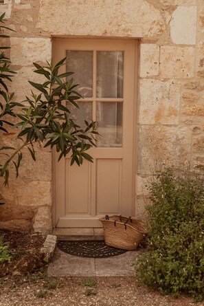 Un bâtiment en pierre avec une porte de couleur beige, un petit marchepied menant à l’entrée et un panier tressé posé sur le sol.