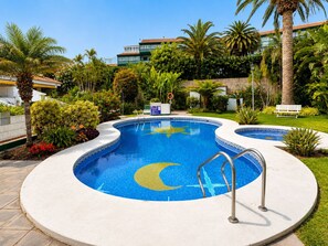 En pool med design som en halvmåne, omgivet af palmer og grønt, med bygninger i baggrunden.