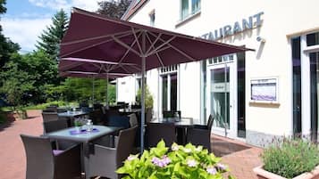 Ein Sitzbereich im Freien mit Tischen und Stühlen, einem großen Sonnenschirm und einem Gebäude mit der Aufschrift 'RESTAURANT'.