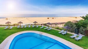 Ein Schwimmbecken mit Liegestühlen und Sonnenschirmen, im Hintergrund ein Strand und ein klarer Himmel.