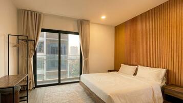 Quarto de hotel com cama, escrivaninha, cadeira, guarda-roupa e vista da cidade através de grandes janelas.