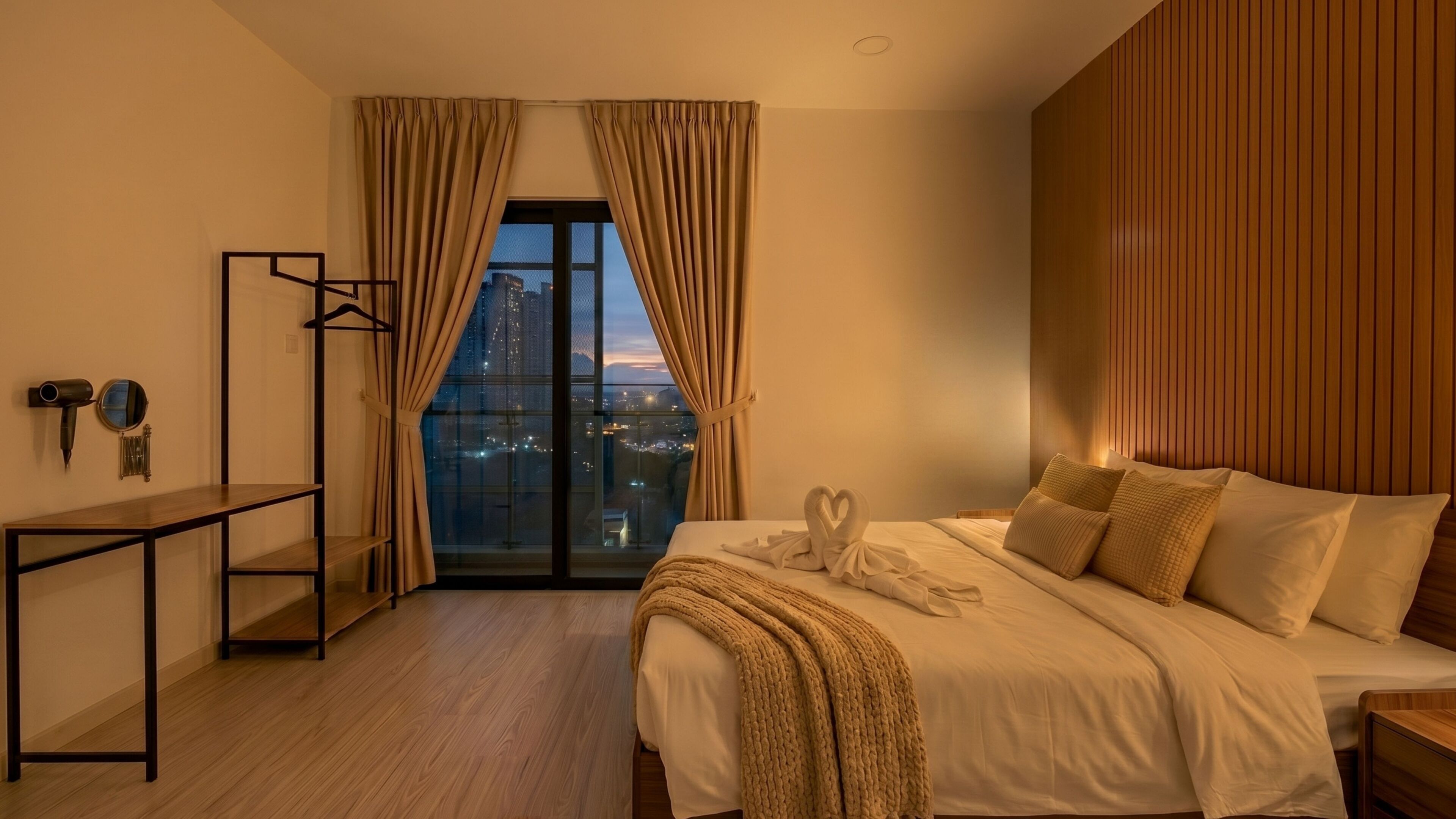 Une chambre d’hôtel comprenant un lit, une tête de lit en bois, une table de chevet, un miroir, un porte-manteau et une vue sur le paysage urbain, visible à travers une grande fenêtre munie de rideaux.
