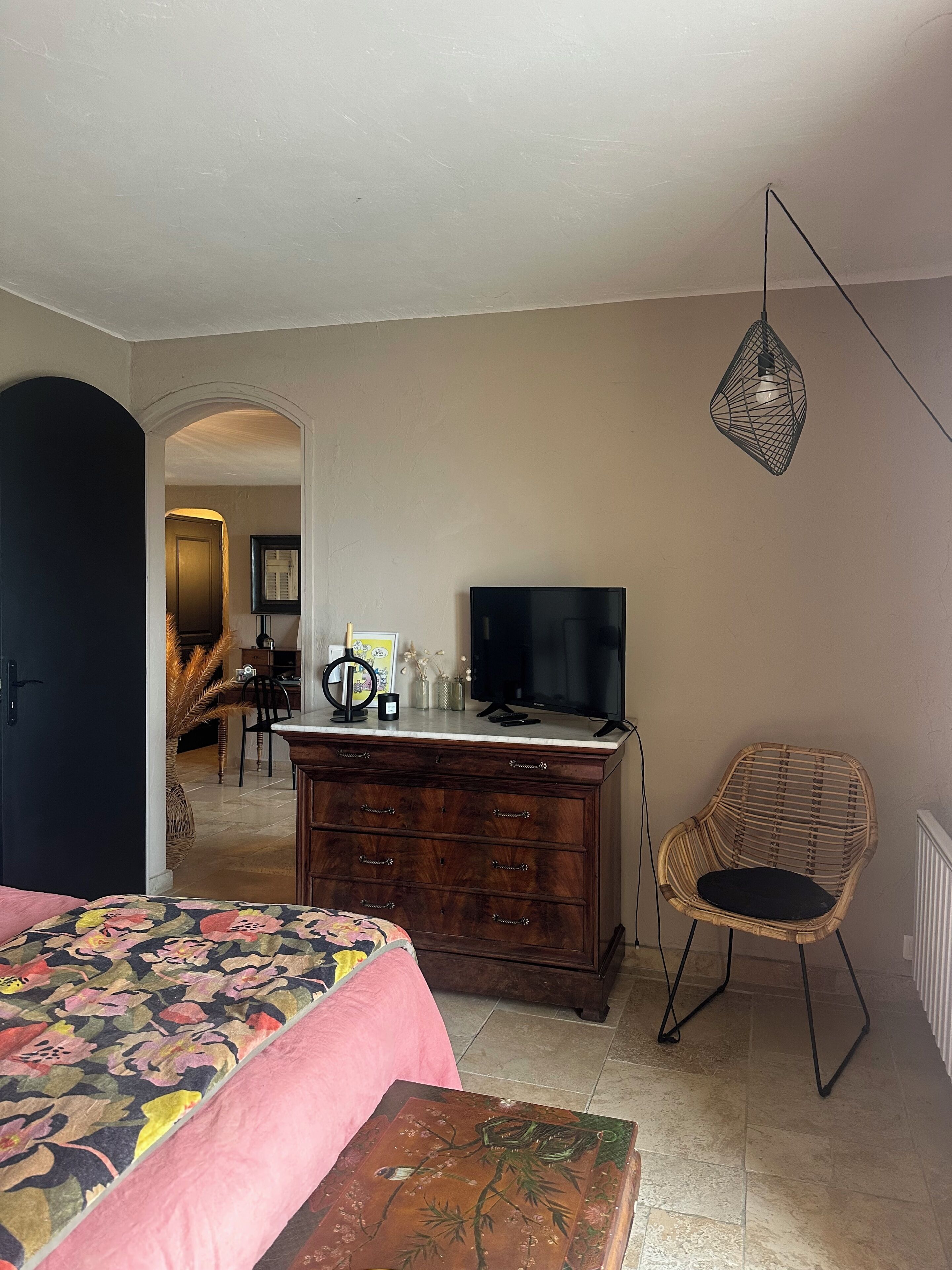 A bedroom with a bed, dresser, TV, chair, and a doorway leading to another room.