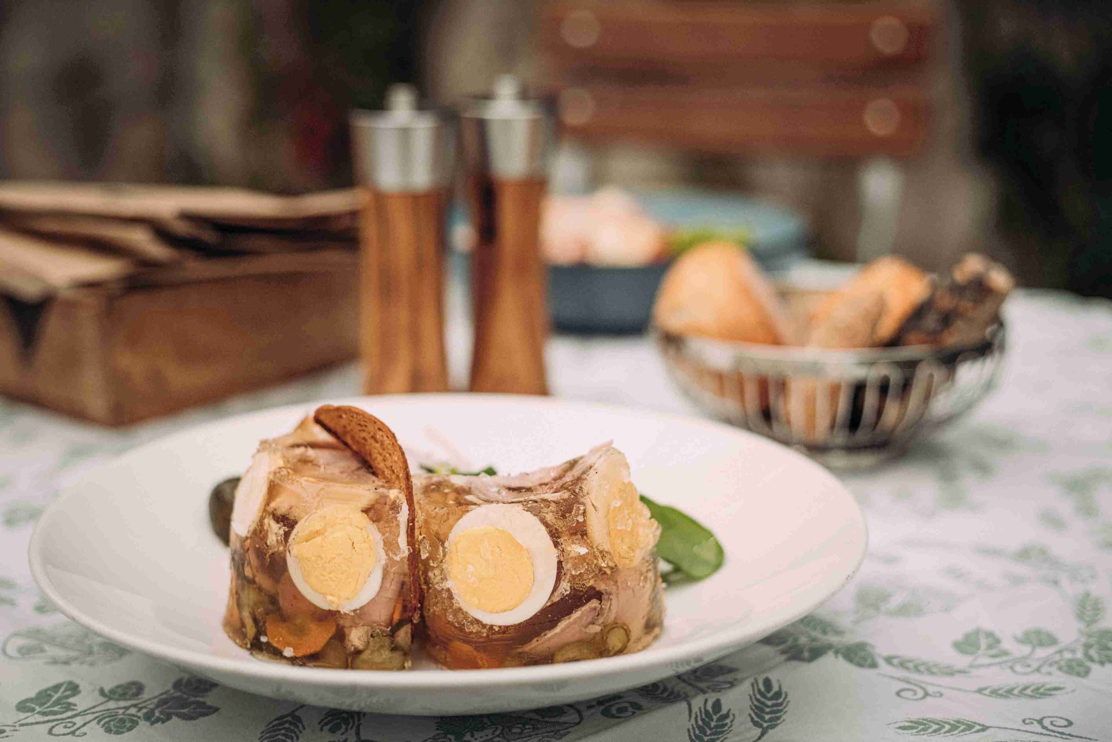 A plate with a rolled meat dish, a bowl of bread, and two salt shakers on a tablecloth.