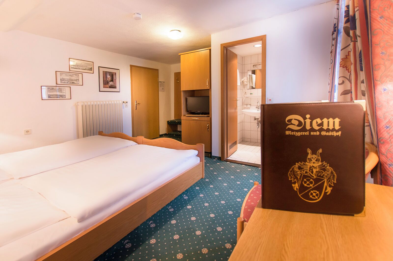Habitación de hotel con cama, un escritorio con un libro, televisión y baño a la vista a través de una puerta abierta.