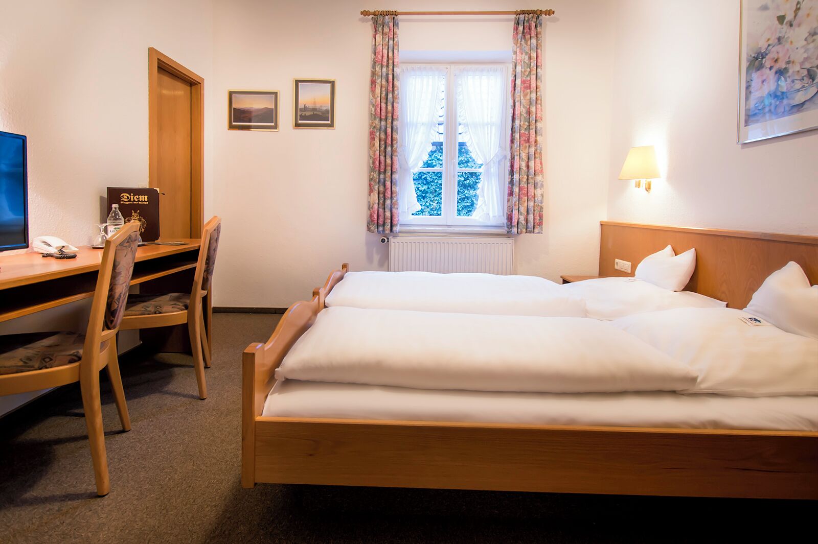 A hotel room with a wooden bed, a desk with a chair, a window with curtains, and a lamp.