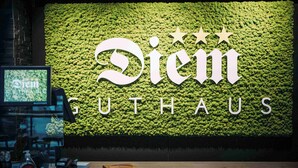 En skylt med texten ”DIEM GUTHAUS” i stora bokstäver, placerad mot en grön bakgrund av lövverk.