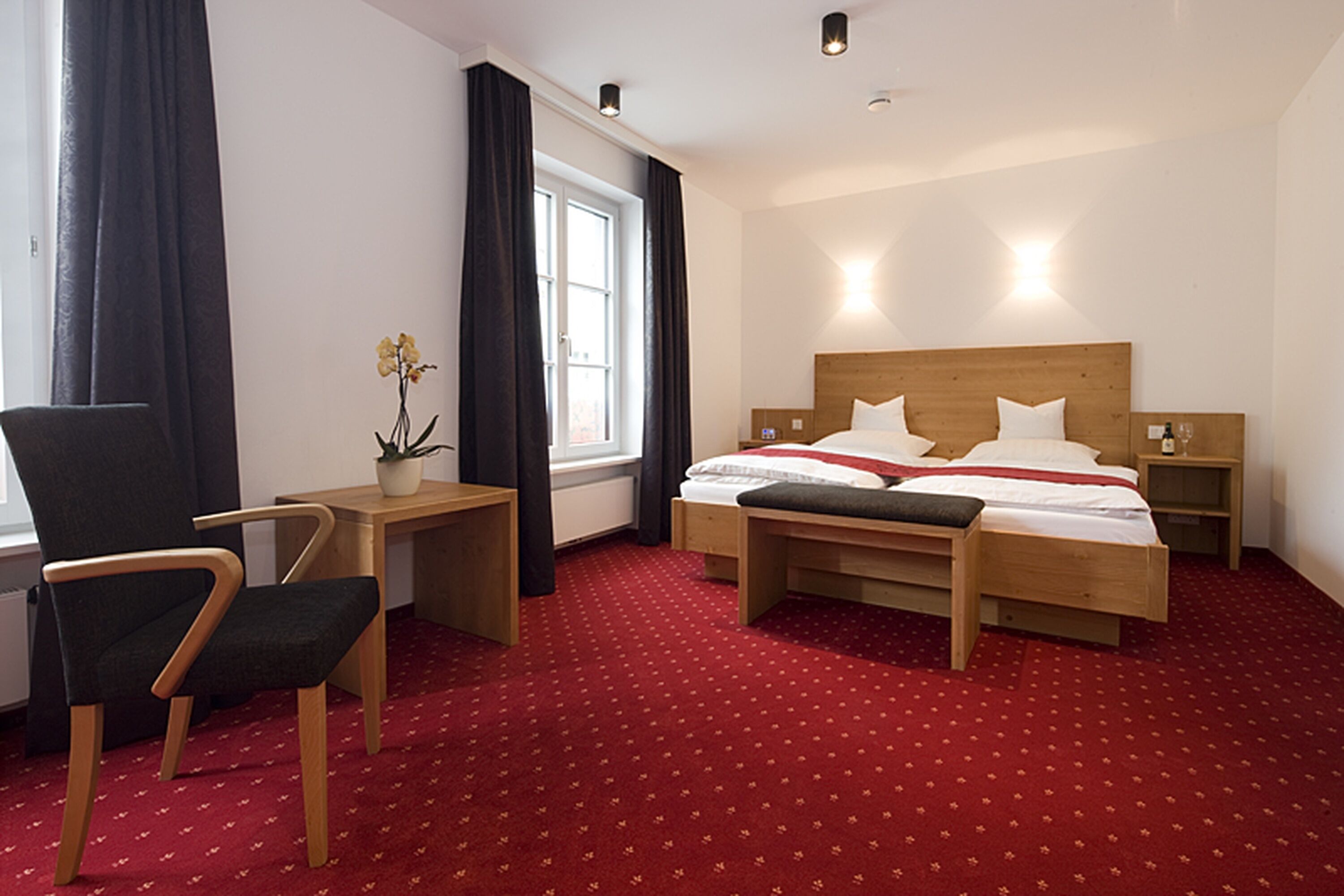 Quarto de hotel com cama, mesas de cabeceira, uma cadeira, janela com cortinas e um vaso com planta.