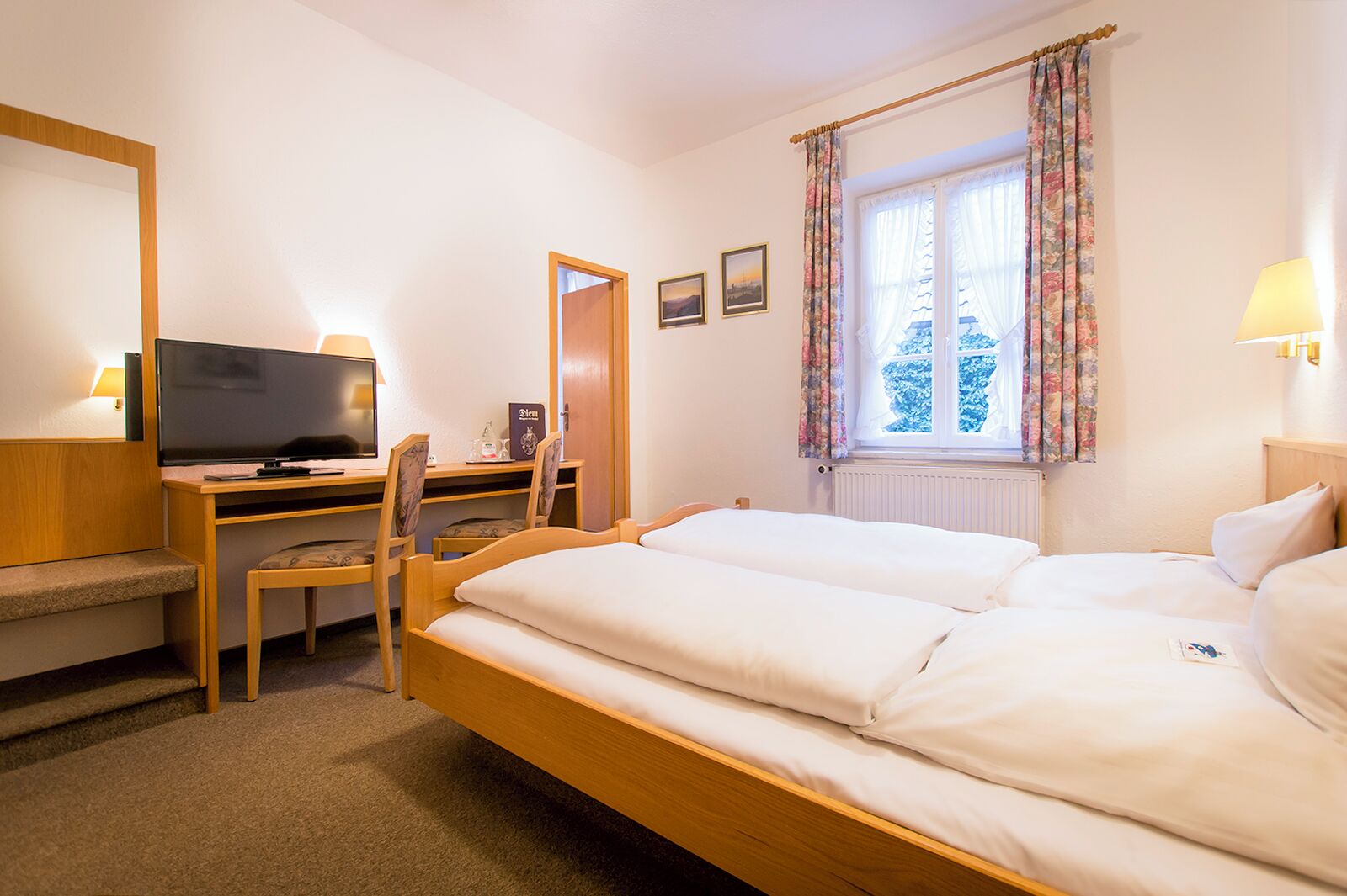 A hotel room with a bed, a desk with a television, a chair, and a window with curtains.