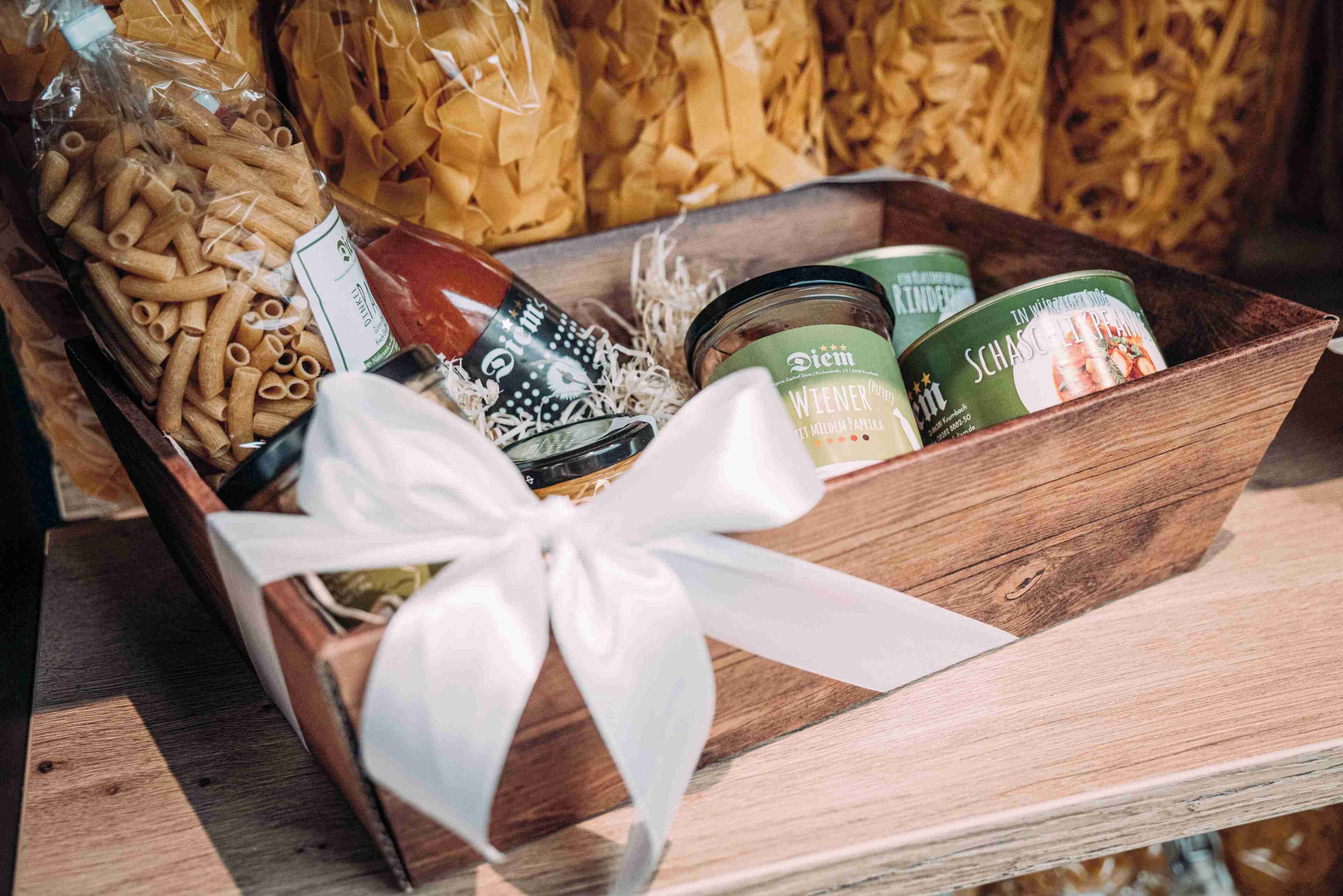 A wooden box with a white ribbon, containing pasta, jars, and a bottle.
