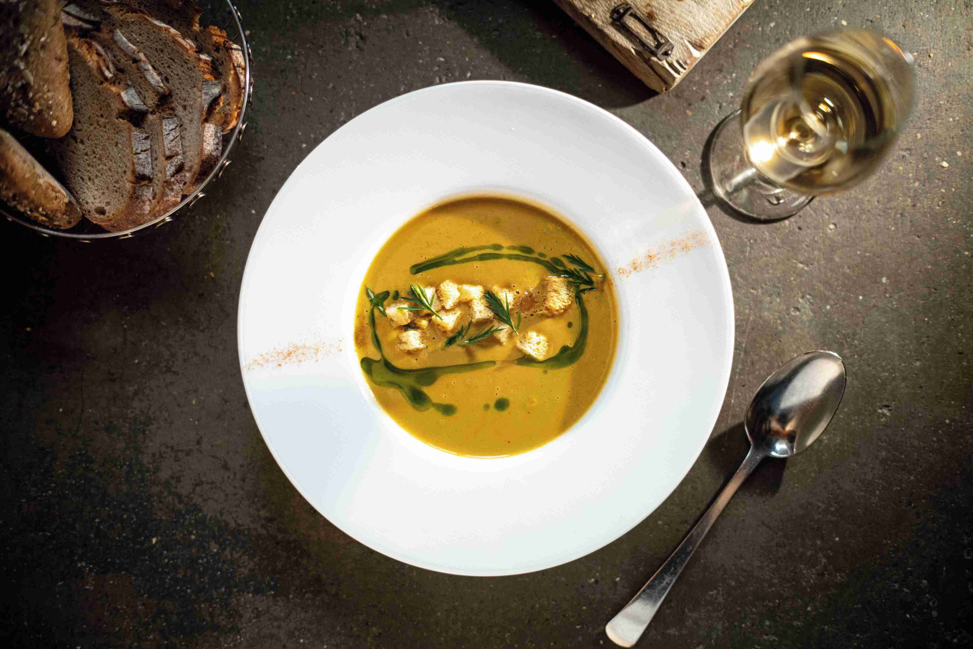 Un plato blanco con sopa adornada con hierbas, una cuchara y una copa de vino blanco sobre una superficie oscura.