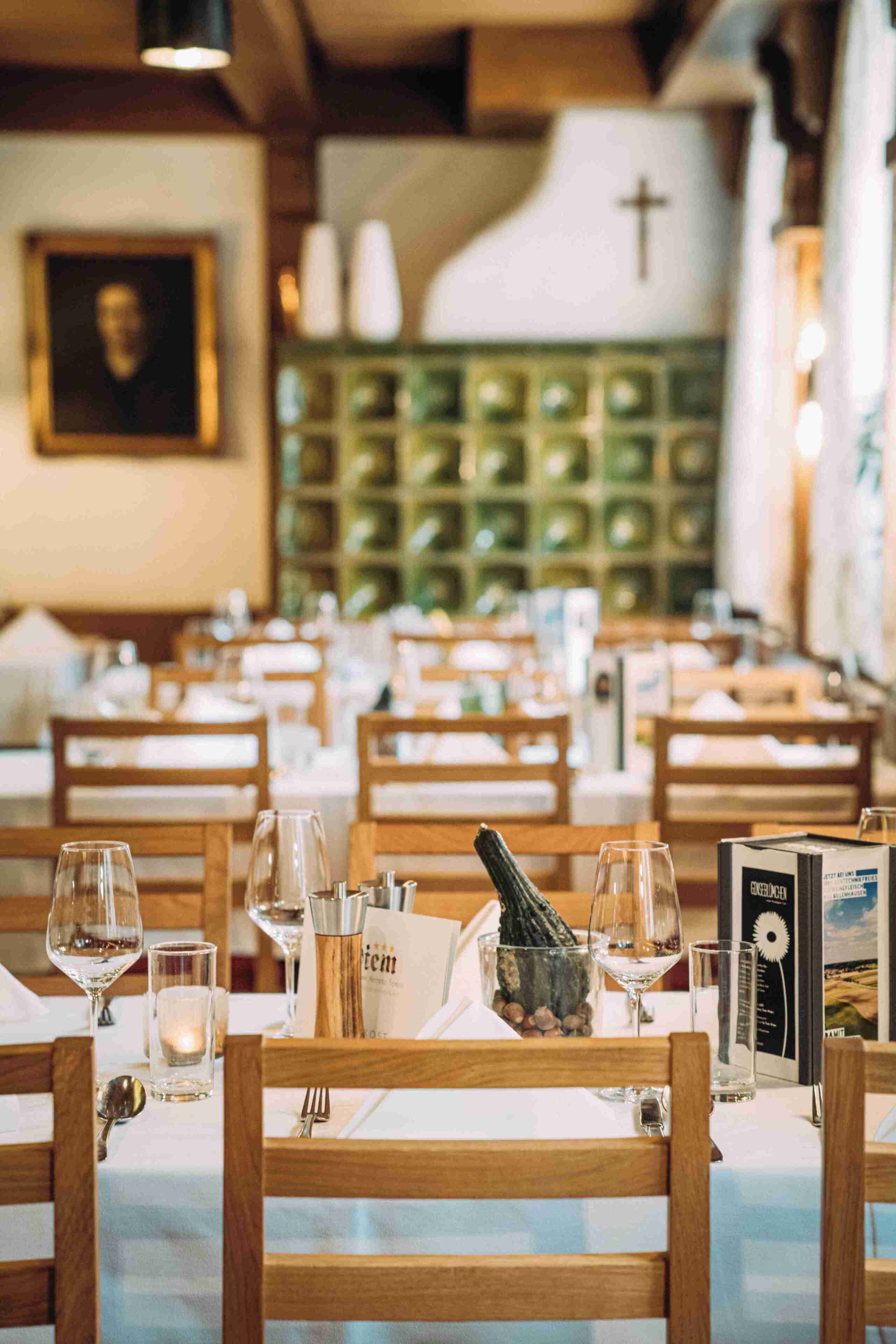Een restaurantinterieur met houten stoelen, tafels gedekt met wijnglazen, een menukaart en een kruis aan de muur.