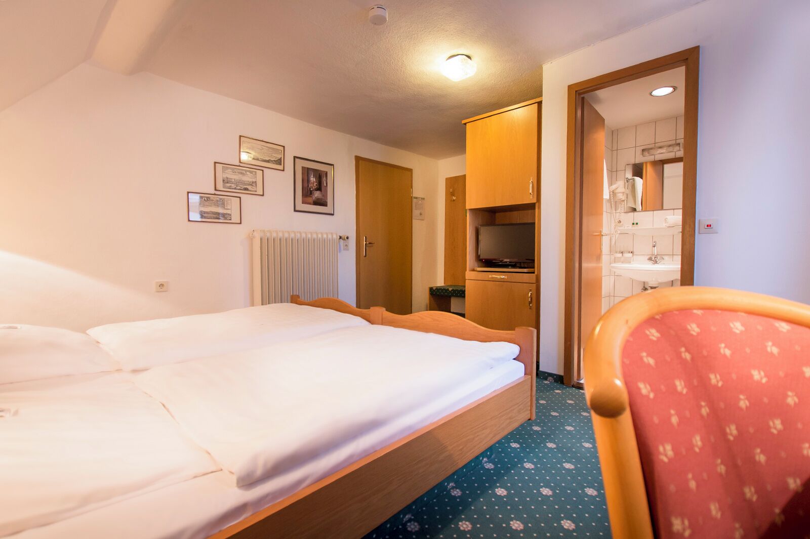 Habitación de hotel con cama doble, armario de madera, televisor y baño con lavamanos y espejo.