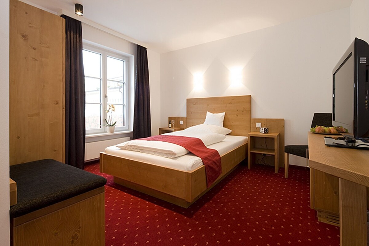 Une chambre d’hôtel avec un lit en bois, un bureau avec un ordinateur, une télévision et une fenêtre avec des rideaux.