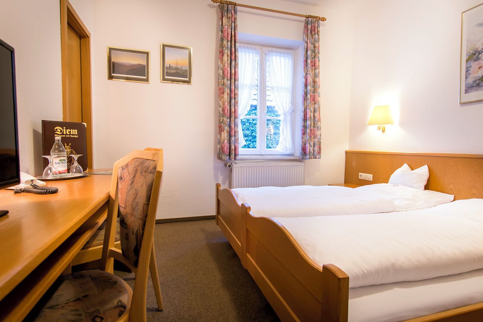 A hotel room with a wooden bed, a desk with a computer, a chair, two framed pictures on the wall, and a window with floral curtains.