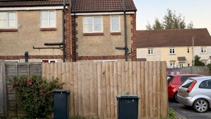 A residential area with two houses, one with solar panels, a wooden fence, and two black trash bins in the foreground. - Newly Renovated 2-Bed House w/ Parking – Sleeps 5 Near Shaftesbury Town (Shaftesbury)