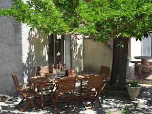Un espace de restauration en plein air avec une table ronde en bois et des chaises, entouré d’un arbre et d’un banc en pierre.