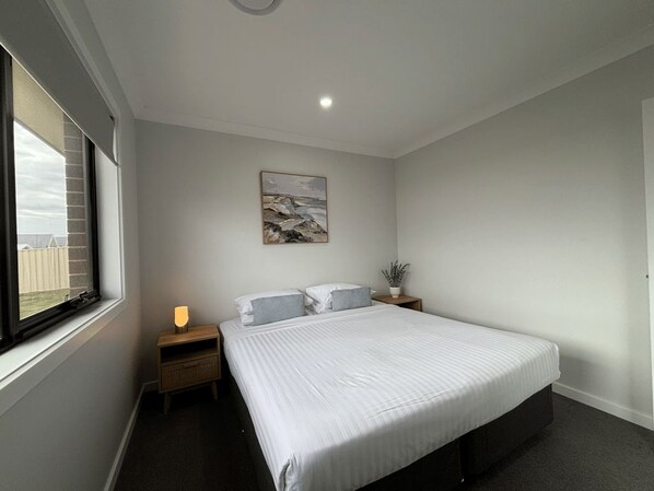 A bedroom with a large bed, a bedside table with a lamp, a wall-mounted painting, and a window with a view. - Wyoming Getaway Two - Sleeps 8 (Wagga Wagga)