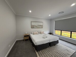 A modern bedroom with a large bed, a nightstand, a wall-mounted artwork, and a window with a view. - Wyoming Getaway Two - Sleeps 8 (Wagga Wagga)