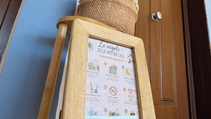 A framed sign with house rules on a wooden stand, a potted plant, and a wooden door. - Fiumicino sea view|Relax&Comfort Near the airport and Fiera di Roma (Isola Sacra)