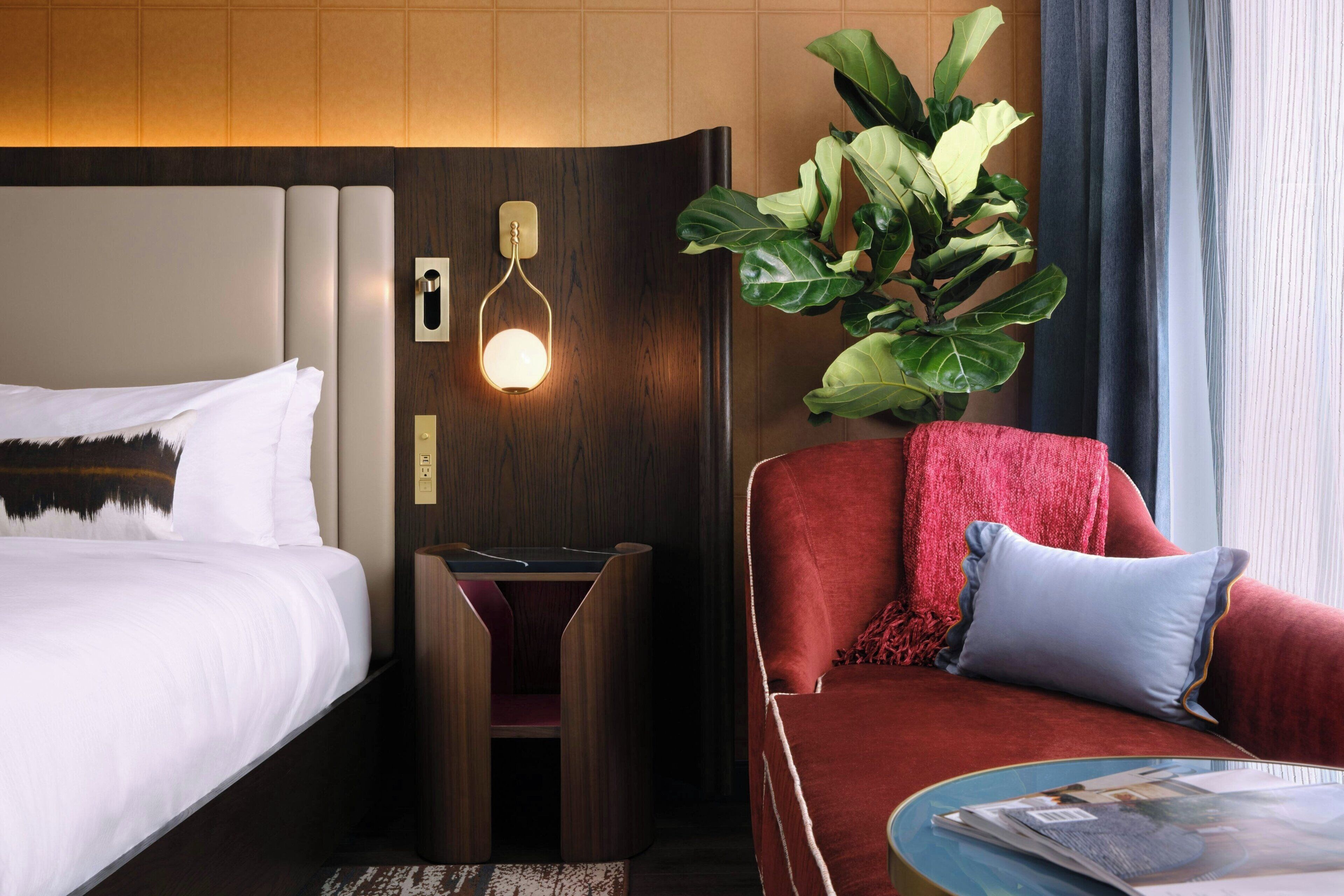A hotel room with a bed, a desk, a red armchair, a large plant, and a window with curtains.