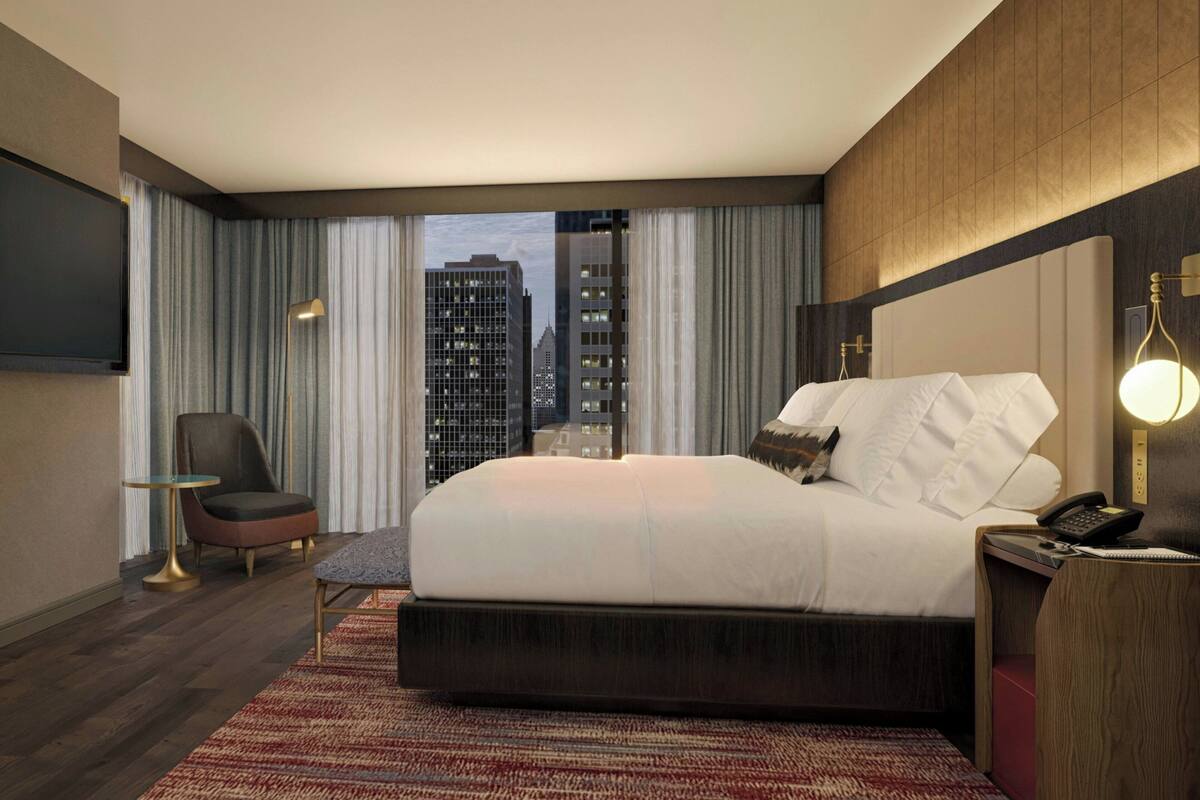 A hotel room with a large bed, a chair, a desk, a TV, and a view of city buildings.
