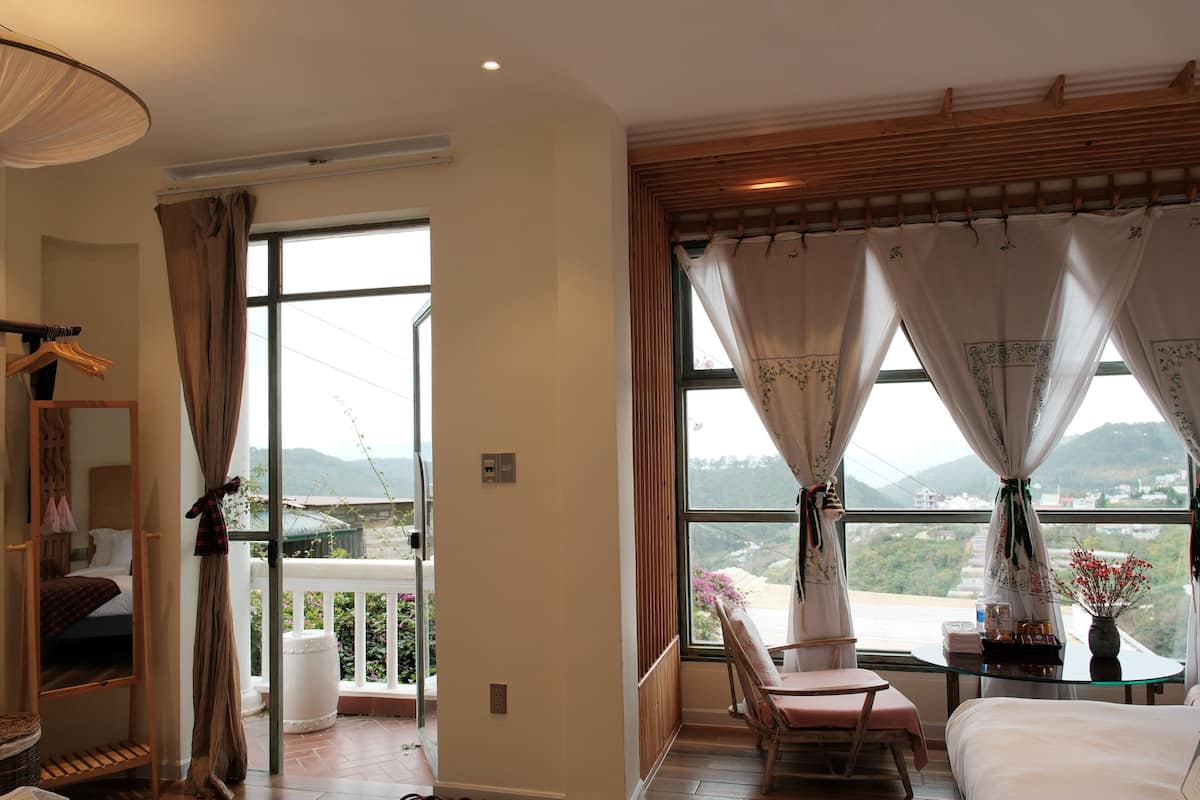 A hotel room with a bed, a chair, a table with a vase, and large windows with curtains.