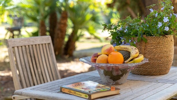 A wooden table with a glass bowl of fruit, a book, and a woven basket with flowers. - Villa del Conte | Exclusive Villa with Private Pool & Panoramic Garden (Alghero)