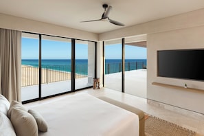 A hotel room with a large bed, a TV, and a balcony with a view of the beach and ocean.
