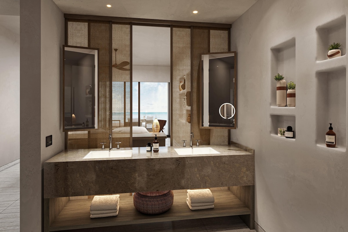 Une salle de bain moderne avec deux lavabos, un grand miroir et une vue sur une chambre à coucher avec un lit et un luminaire suspendu.