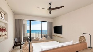 Quarto de hotel com uma cama grande, televisão, ventoinha no teto, vista para a praia e um espaço de estar também com vista.