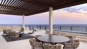 Uma área de estar coberta ao ar livre com vista para o oceano, apresentando uma mesa redonda e cadeiras de vime.