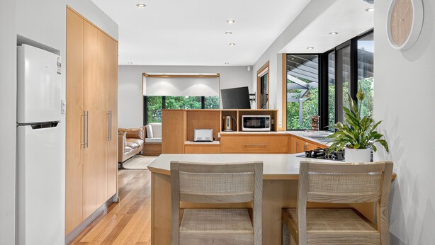 A modern kitchen with wooden cabinets, a white refrigerator, a dining table with two chairs, and a large window offering a view of greenery. - Luxury Coastal Escape at Kingston Beach (Kingston Beach)