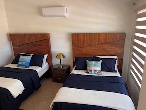 Dos camas con cabeceros de madera, una mesita de noche con lámpara y un equipo de aire acondicionado en la pared.