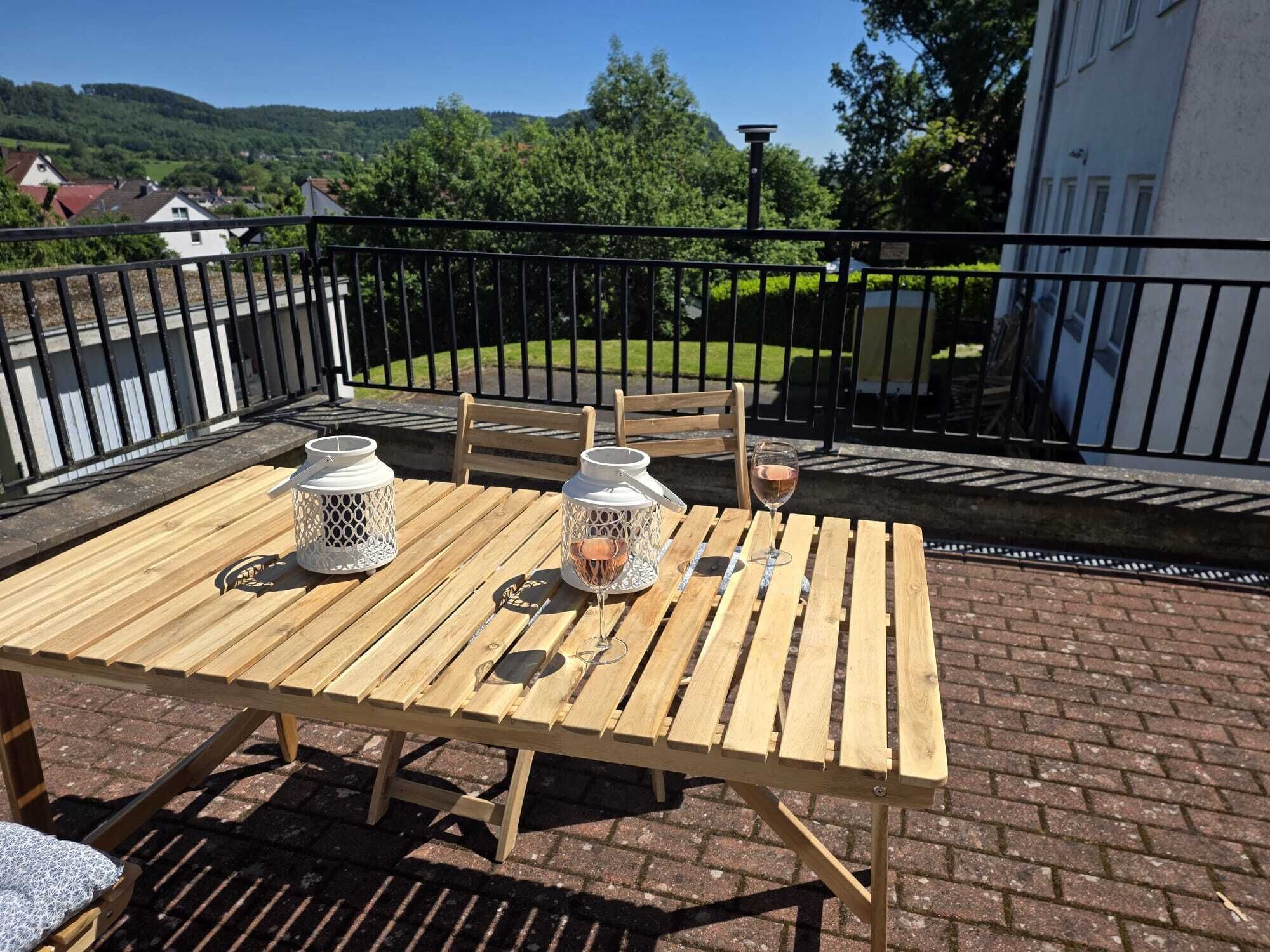 Un tavolo di legno con due lanterne bianche e un bicchiere di vino su un balcone con pavimento in mattoni e vista su un paesaggio verde.