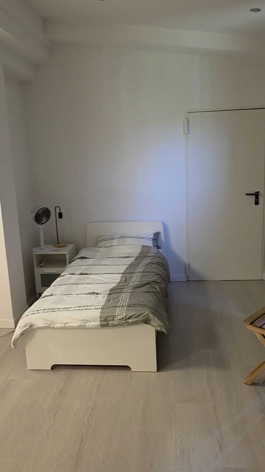 Un dormitorio con una cama, una mesita de noche con un ventilador, una puerta y un taburete de madera.