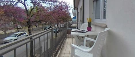 Un balcón con una mesa y sillas blancas, una planta en maceta y vistas a una calle con coches y árboles.