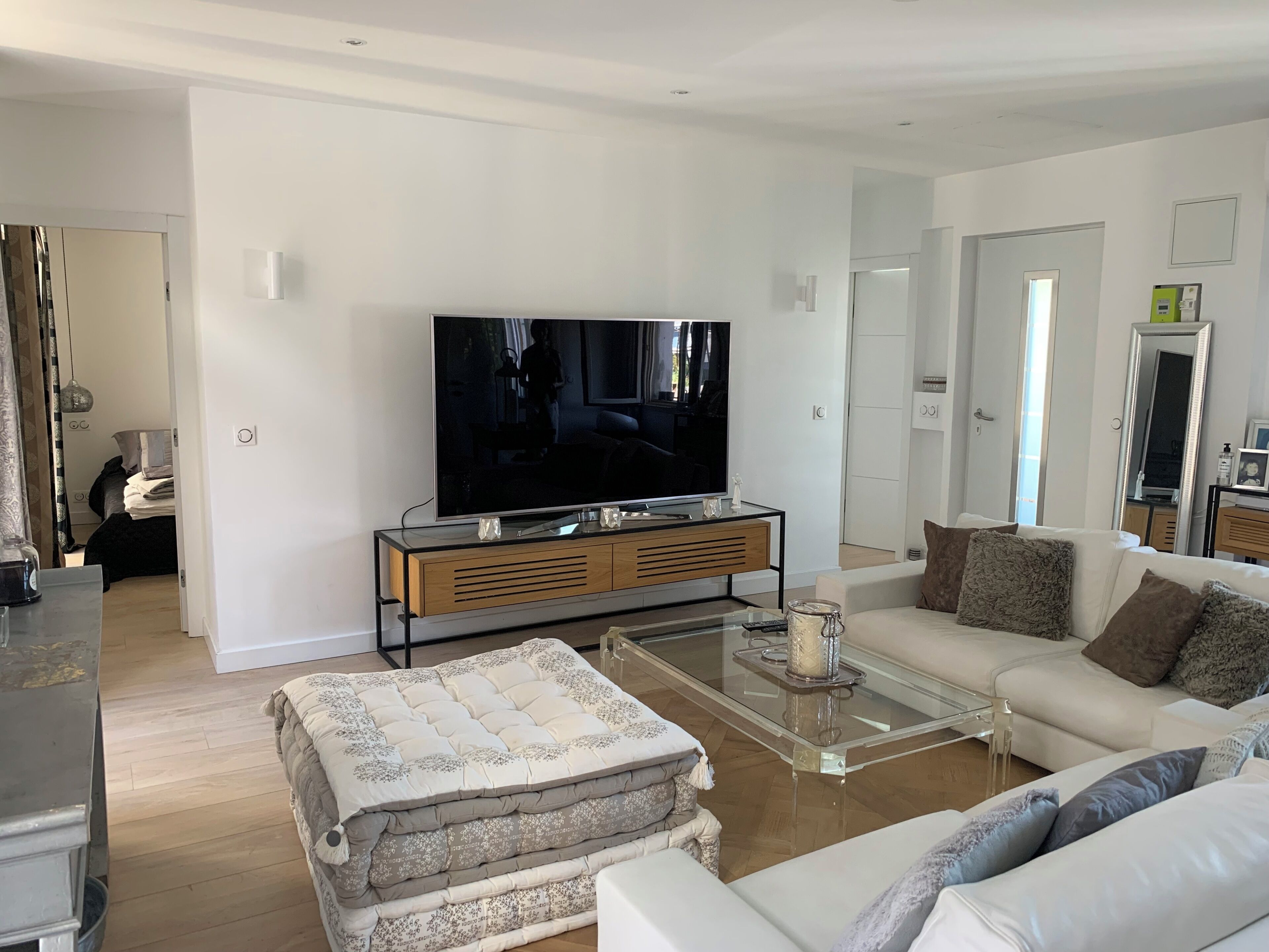A modern living room with a flat-screen TV, a glass coffee table, a sofa, and a bed with a patterned duvet.