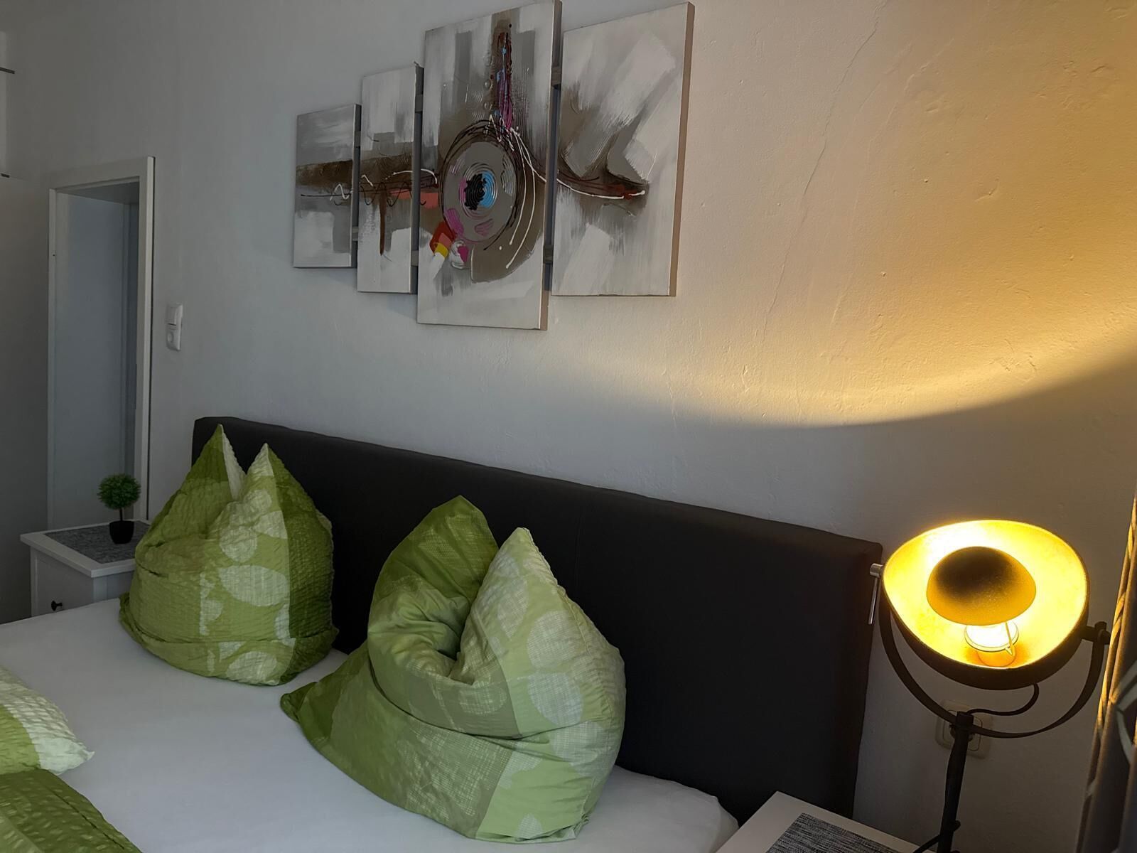 Ein Schlafzimmer mit einem Bett, grünen Kissen, einem Nachttisch mit Lampe und abstrakter Wandkunst.