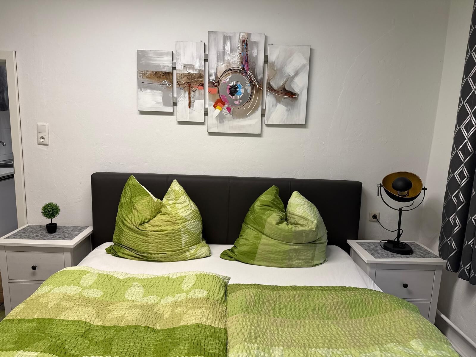 Un dormitorio con una cama, almohadas verdes, una mesita de noche con lámpara, un reloj de pared y arte abstracto.