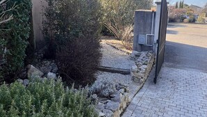 Une allée pavée mène à une entrée fermée par un portail, l’ensemble étant entouré de divers arbustes et d’un mur de pierres.