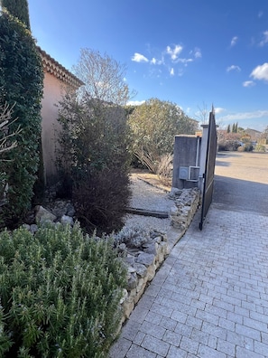 A paved driveway leading to a gated entrance, surrounded by various shrubs and a stone wall.