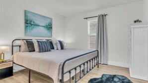 A bedroom with a bed, a nightstand, a painting, a window with curtains, and a door. - Cozy Retreat on Dana Drive (Haltom City)