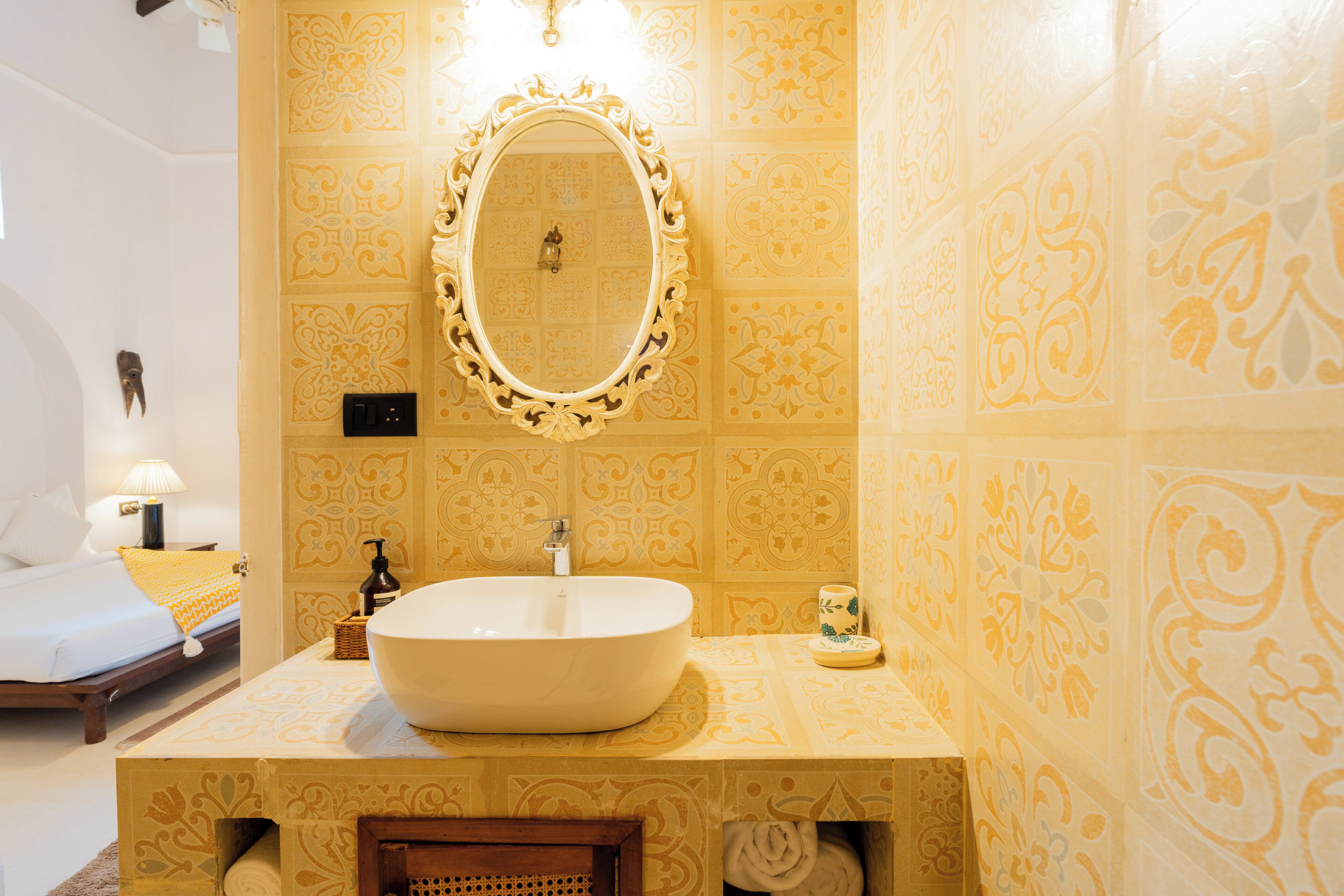 Une salle de bain avec un grand lavabo ovale, un miroir doté d’un cadre orné, des carreaux muraux à motifs, et un lit avec un oreiller jaune et une table de chevet avec une lampe.