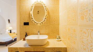 Une salle de bain avec un grand lavabo ovale, un miroir doté d’un cadre orné, des carreaux muraux à motifs, et un lit avec un oreiller jaune et une table de chevet avec une lampe.
