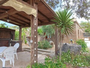 Un espace extérieur abrité, doté d’une pergola en bois, d’un mur en pierre et d’une table en plastique blanche avec des chaises.