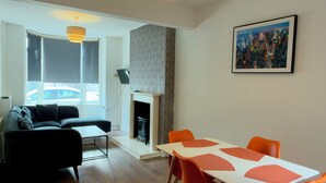 Een moderne woonkamer met een open haard, een eethoek met oranje stoelen en een groot raam met jaloezieën.