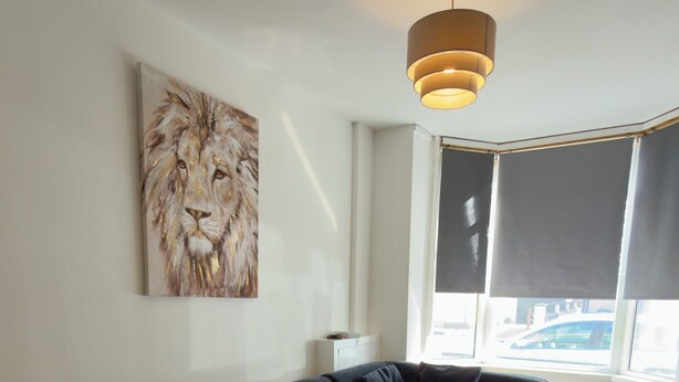 Een moderne woonkamer met een zwart hoekbank, een kleine witte salontafel, een plafondlamp en een groot raam met jaloezieën.